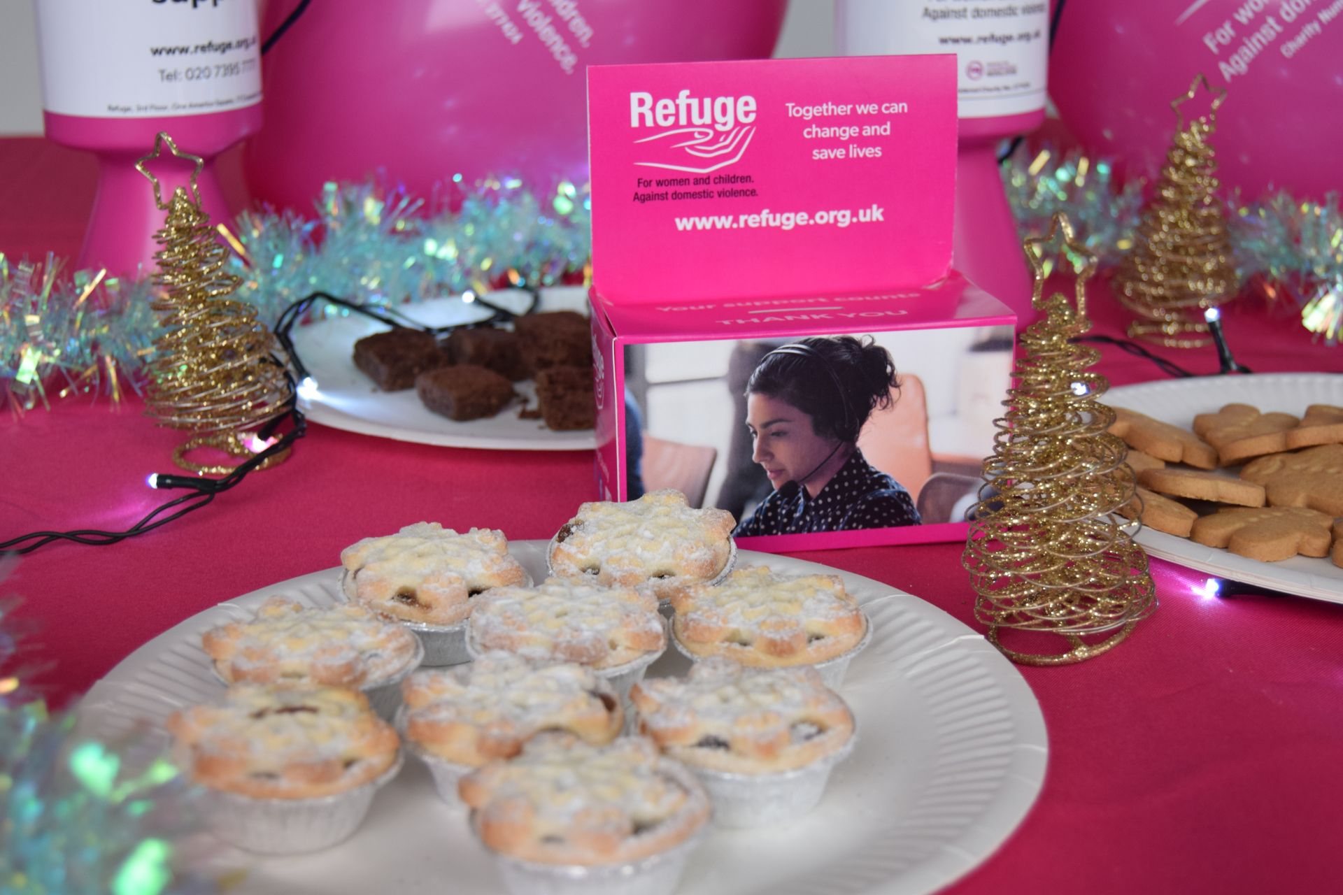 A festive bake sale table with mince pies, biscuits, and a Refuge charity donation box.