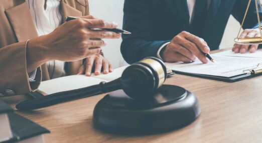 Gavel resting in a courtroom. Behind, a man and a woman's hands can be seen holding pens over legal documents.