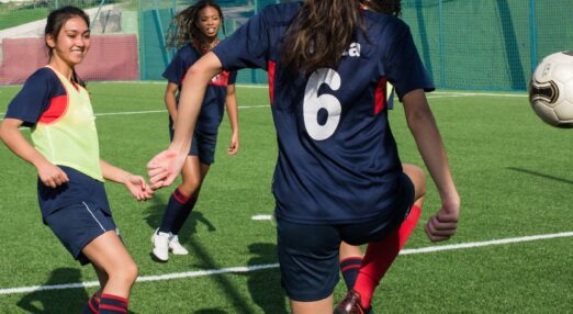 A young woman, facing away from camera, is on a football pitch wearing a navy football kit. The ball is in the air to her right, and two other women in football kit are running towards it.