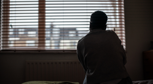 Back of a woman sat on a bed, silhouetted against window with blinds drawn